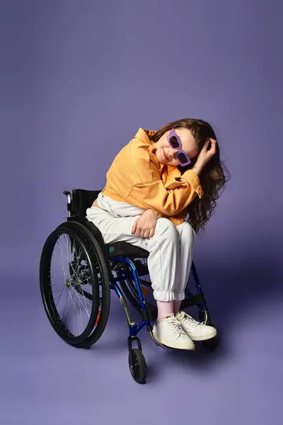 A young woman in a wheelchair, sporting sunglasses and casual attire, sits in a studio with a purple backdrop. — Stock Photo