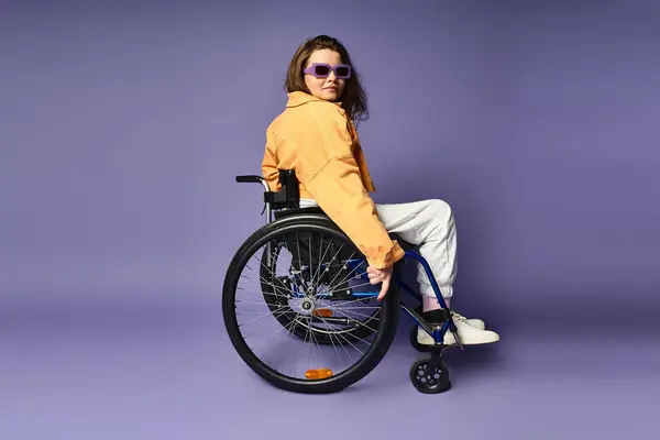 A young woman in a wheelchair and fashionable casual attire poses against a purple backdrop. — Stock Photo