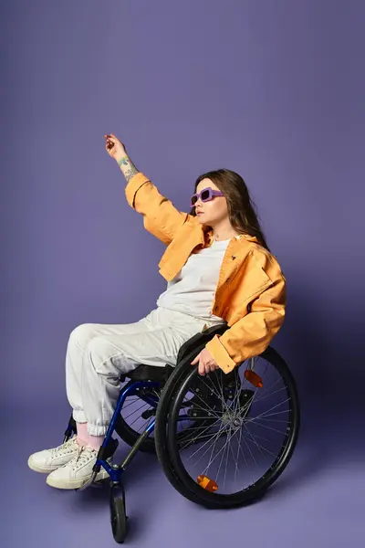 A young woman in a yellow jacket and white pants sits in a wheelchair in front of a purple background, her arm raised in a pose. — Stock Photo