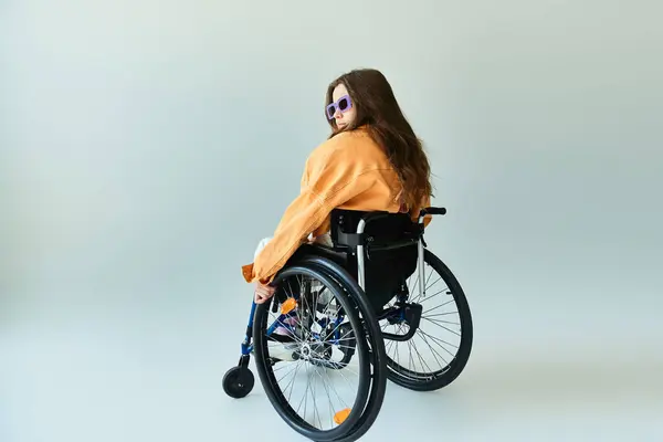 Une jeune femme en fauteuil roulant, vêtue de vêtements décontractés et de lunettes de soleil, pose en studio. — Photo de stock