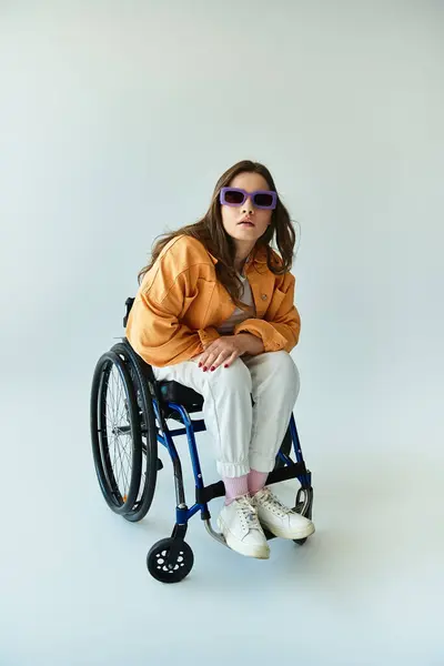 A young woman with long brunette hair sits in a wheelchair and looks at the camera while wearing sunglasses and casual attire. — Stock Photo