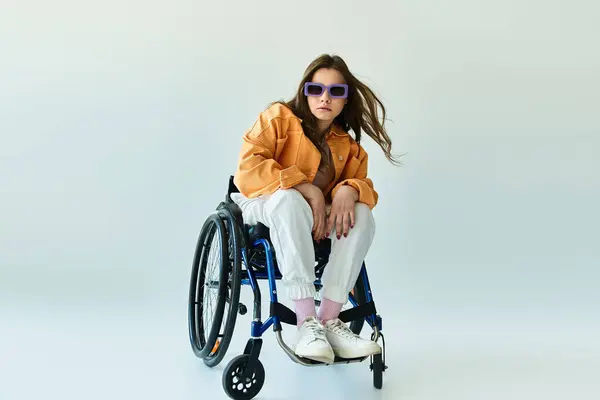 A brunette woman wearing a yellow jacket and white pants sits in a wheelchair in a studio setting with a gray backdrop. — Stock Photo