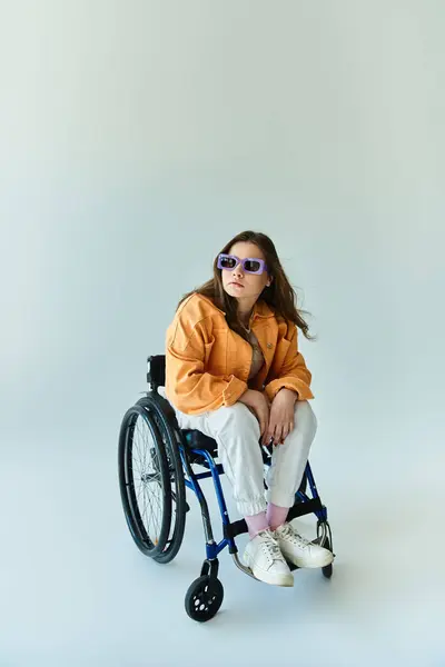 Une jeune femme élégante en fauteuil roulant pose dans un studio sur un fond gris, portant une veste orange vif et des lunettes de soleil. — Photo de stock