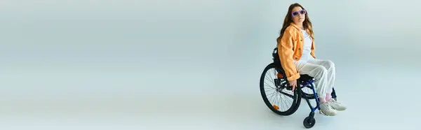 Una joven en silla de ruedas y elegante atuendo casual posa en un estudio con un fondo gris. — Stock Photo