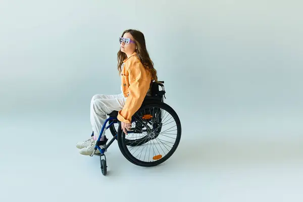 A young woman sits in a wheelchair in a studio with a grey background. She is wearing casual clothing and sunglasses. — Stock Photo
