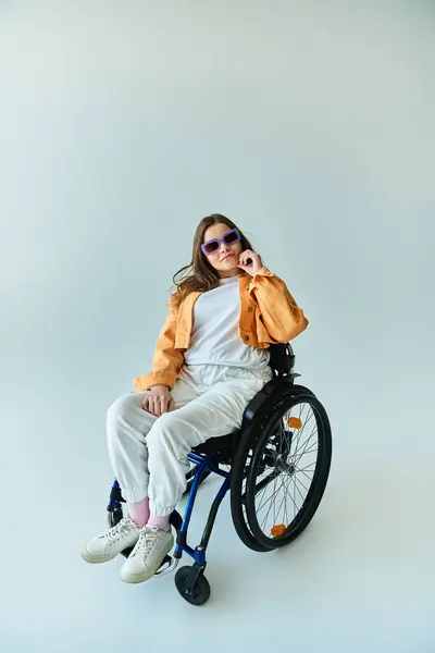 Una joven morena con estilo en gafas de sol se sienta en una silla de ruedas posando para una foto en un estudio con un fondo gris - foto de stock