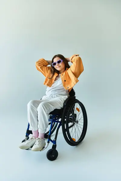 Jeune femme en fauteuil roulant porte chemise blanche, veste orange, lunettes de soleil, mains sur la chaise, regardant la caméra en studio — Photo de stock