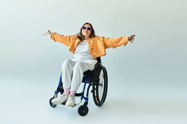 Eine junge Frau im Rollstuhl lächelt, als sie ihre Arme ausstreckt, in stylischer Freizeitkleidung und Sonnenbrille. — Stockfoto