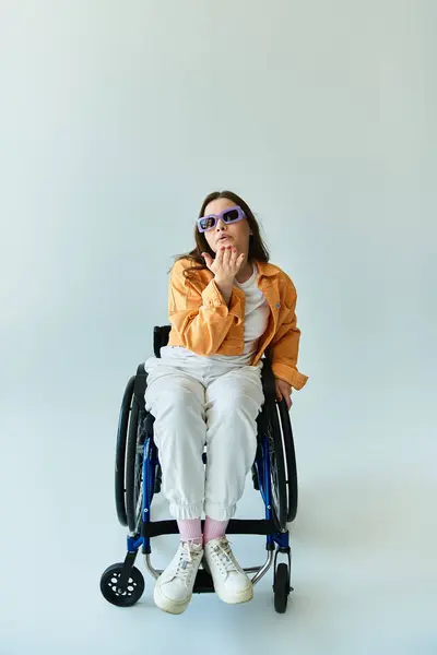 A young woman in a wheelchair, wearing sunglasses and a casual outfit, sits in a studio with a gray background. — Stock Photo