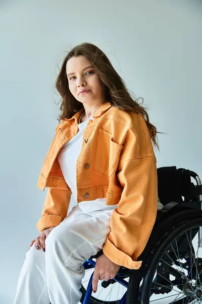 A young brunette woman wearing a white t-shirt and orange denim jacket poses in a wheelchair against a grey background. — Stock Photo