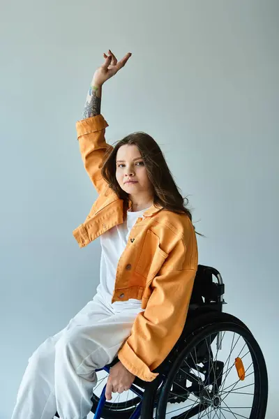 A brunette woman in a wheelchair sits in a studio, wearing a stylish orange jacket and white pants, with one arm raised. — Stock Photo