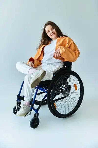 A young brunette woman sits in a wheelchair, wearing a stylish orange jacket and white pants, smiling at the camera in a studio setting. — Stock Photo