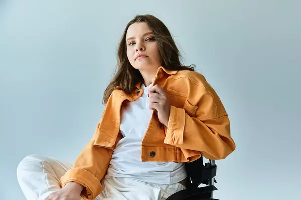 A young brunette woman sits in a wheelchair against a grey backdrop, wearing stylish casual clothes and adjusting her orange jacket. — Stock Photo