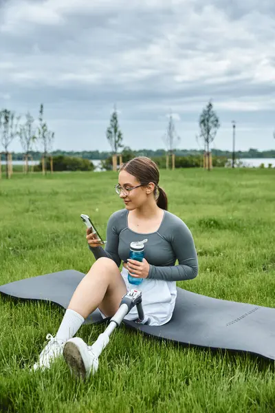 A young woman with a prosthetic leg relaxes on a mat while checking her phone in a lush green park. — Stock Photo