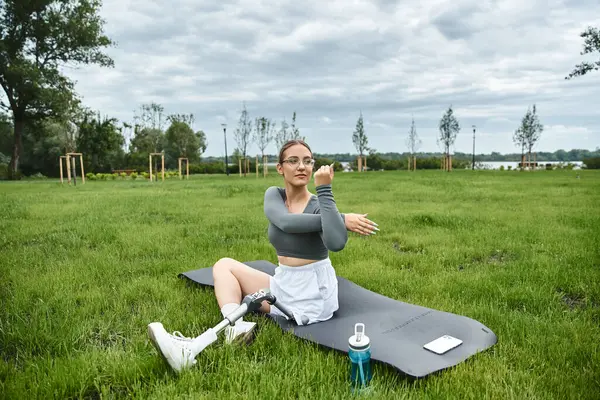 Una mujer decidida se ejercita sobre una alfombra en una exuberante vegetación, estirando su brazo mientras las nubes se ciernen sobre la cabeza. - foto de stock