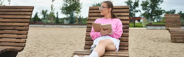 Uma jovem no sportswear relaxa, lendo um livro em uma praia de areia, abraçando o ar livre com alegria. — Fotografia de Stock