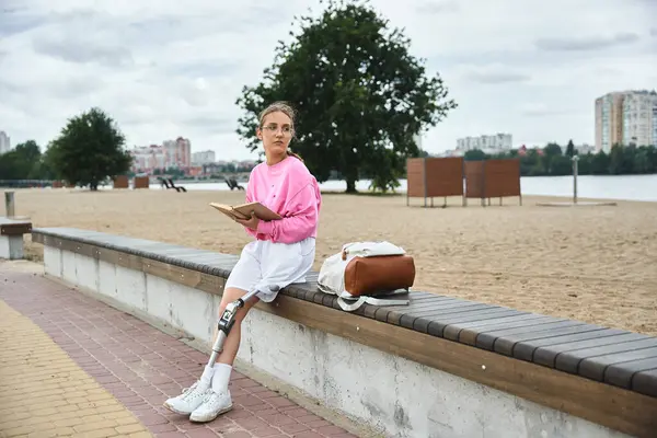 A young woman in sportswear embraces the outdoors, reading by the beach with her prosthetic leg. — Stock Photo