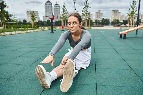 A young woman works out in sportswear, stretching her legs on a green fitness surface in an urban park. — Stock Photo