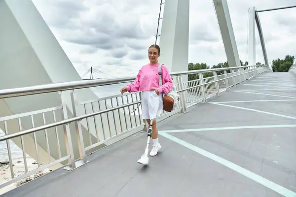 A spirited young woman in sportswear confidently walks across a modern bridge, enjoying her day outdoors. — Stock Photo