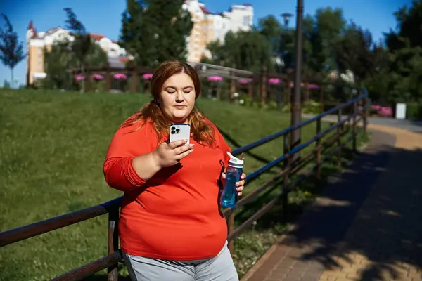 Una mujer de talla grande comprueba su teléfono mientras hace ejercicio al aire libre en un parque vibrante. — Stock Photo