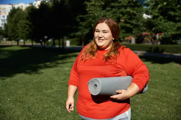 A confident woman smiles while holding a yoga mat in a vibrant green park. — Stock Photo