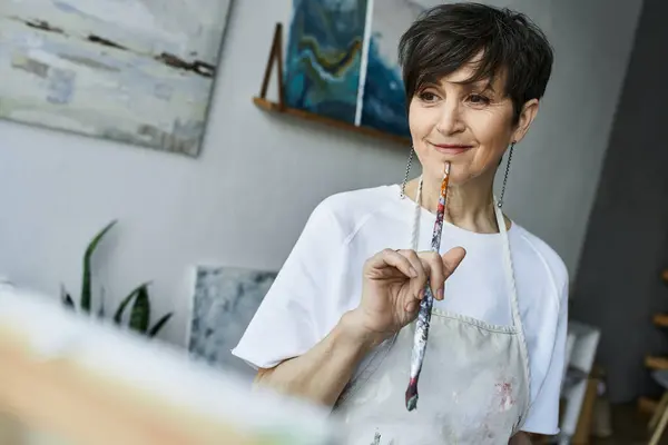 A woman reflects thoughtfully while holding a paintbrush in her studio. — Stock Photo