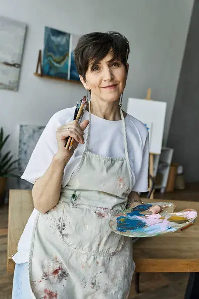 A woman in an art studio smiles while holding brushes and a colorful palette. — Stock Photo