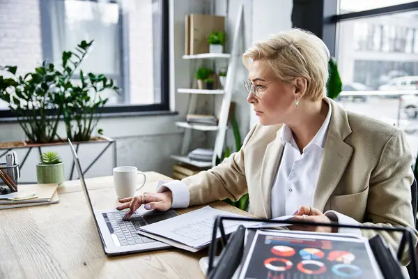 Una donna adulta vestita elegantemente si concentra sul suo computer portatile sorseggiando caffè in uno spazio di lavoro alla moda. — Foto stock