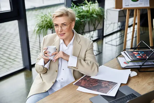A confident woman in chic attire sips coffee at her stylish desk, surrounded by plants. — Stock Photo