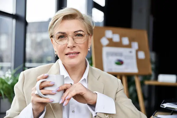 Una donna elegante in abiti eleganti si siede a una scrivania, assaporando il suo caffè in uno spazio di lavoro stimolante. — Foto stock