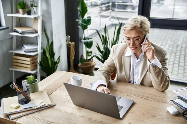 A fashionable woman engages in a phone call while efficiently working on her laptop at a sleek desk. — Stock Photo