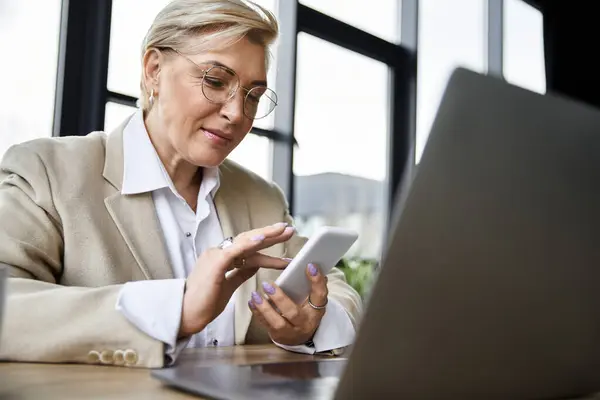 Une femme élégante travaille avec diligence à son bureau, en utilisant son smartphone alors qu'elle examine son ordinateur portable. — Photo de stock