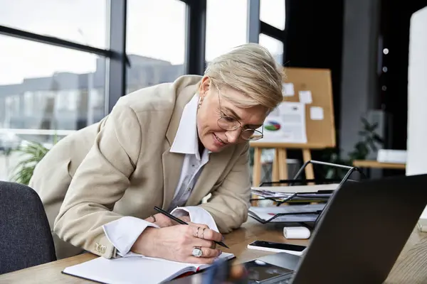 Uma mulher elegante diligentemente escreve notas enquanto se inclina sobre uma mesa em um espaço de trabalho contemporâneo. — Stock Photo