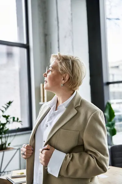 Uma mulher elegante em traje elegante reflete enquanto olha para fora de uma grande janela em seu local de trabalho. — Fotografia de Stock