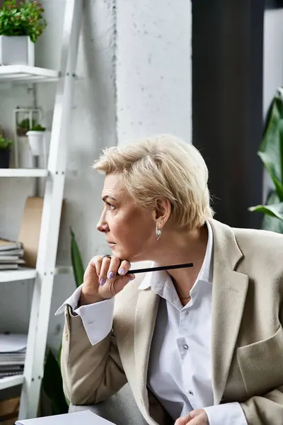 Una mujer adulta vestida con elegancia reflexiona sobre las ideas mientras está sentada en un espacio de trabajo contemporáneo. - foto de stock