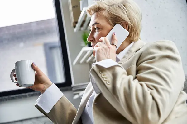 An adult woman in elegant attire enjoys a coffee while engaged in a phone conversation. — Stock Photo
