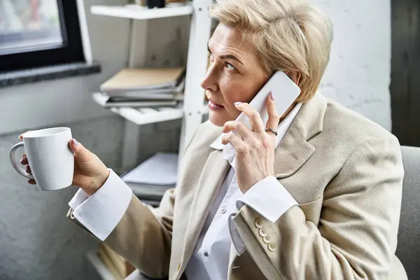An elegant woman is engaged in a phone conversation while sipping coffee in a modern workspace. — Stock Photo