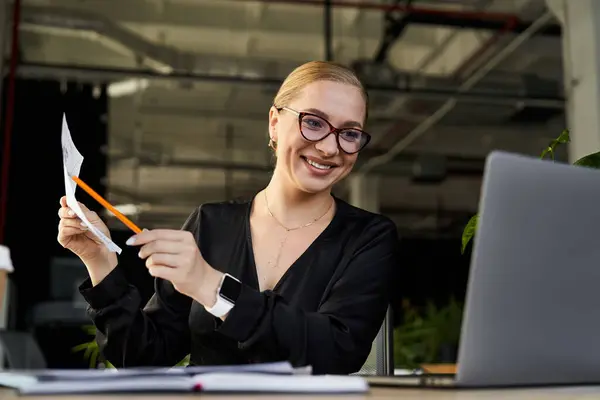 Una giovane donna allegra esamina i documenti mentre si impegna con il suo computer portatile in un ufficio. — Foto stock