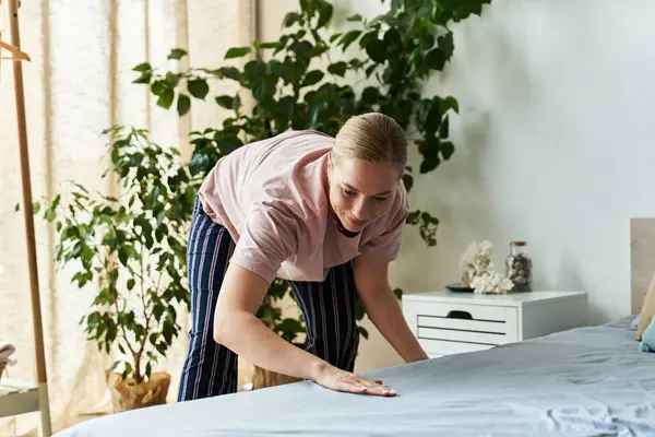 A plus size woman refreshes her bedroom by smoothing the bedspread, surrounded by greenery. — Stock Photo
