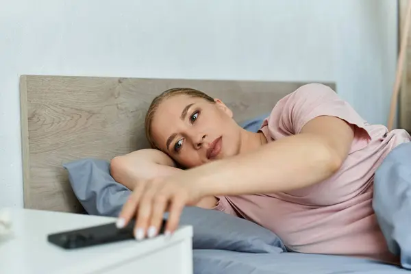 A young plus size woman lounges comfortably in bed, reaching for her remote. — Stock Photo