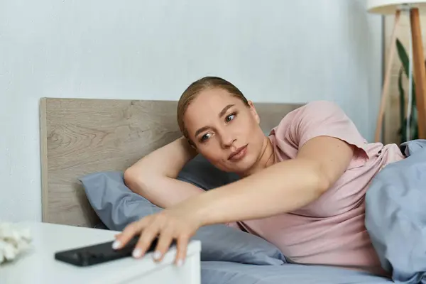 Uma jovem encantadora alcança o controle remoto em sua mesa de cabeceira enquanto relaxa em casa. — Fotografia de Stock