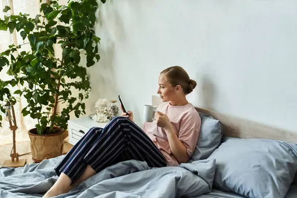A mulher relaxa na cama, tomando café enquanto rola em seu telefone em um ambiente sereno em casa. — Fotografia de Stock