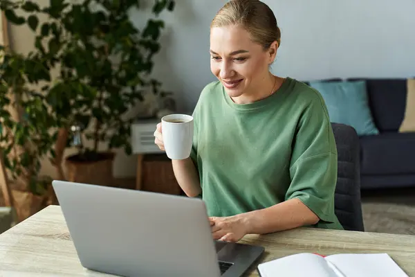 A cheerful young woman holds a mug and types joyfully on her laptop in a bright room. — Stock Photo