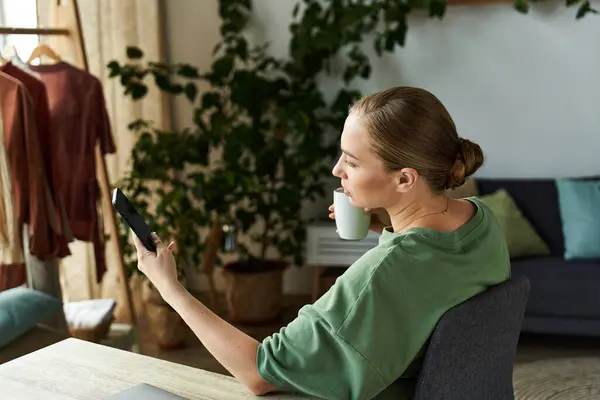 Momento relaxado em casa como uma mulher jovem e bonita desfruta de sua bebida enquanto usa seu telefone. — Stock Photo