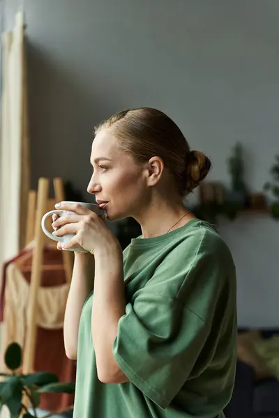 Una bella giovane donna in un accogliente vestito verde assapora il suo caffè a casa, persa nel pensiero. — Foto stock