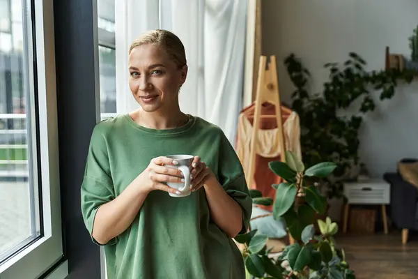 Una joven hermosa mujer de talla grande se para junto a la ventana, saboreando su bebida caliente con comodidad. — Stock Photo