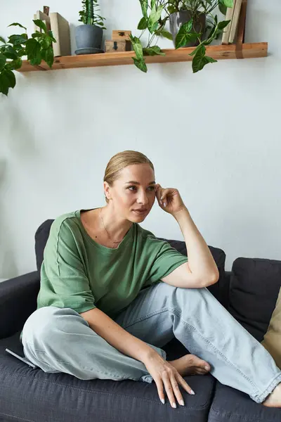 A young plus size woman sits thoughtfully on a sofa, surrounded by cozy houseplants. — Stock Photo