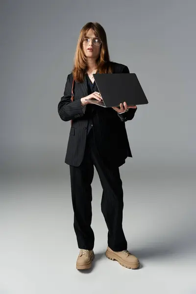 A young man in trendy attire holds a laptop, standing confidently in a sleek studio environment. — Stock Photo