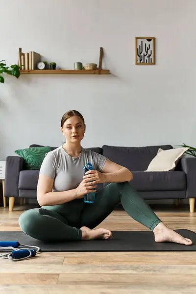 Una joven deportiva se relaja después de un entrenamiento en casa, sosteniendo una botella de agua en su esterilla de yoga. - foto de stock