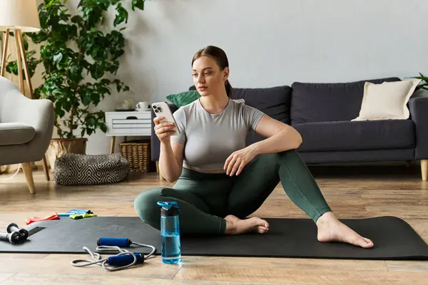 Eine junge Frau in aktiver Kleidung sitzt auf ihrer Yogamatte und checkt ihr Handy in einer gemütlichen häuslichen Umgebung. — Stockfoto